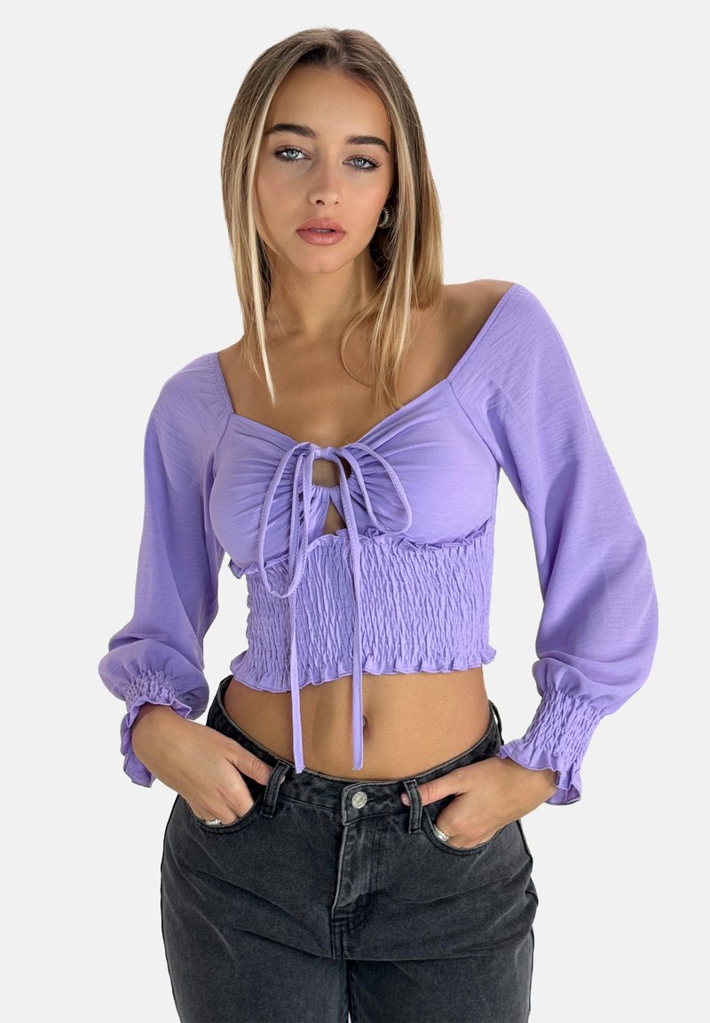 Lila cropped blouse met een smocktaille, strikdetail bij de halslijn en lange pofmouwen, gecombineerd met hooggesneden zwarte jeans.