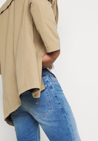 Beige oversize jacka med korta ärmar, bärs över en vit t-shirt, i kombination med blå denimjeans som har en liten röd logotyp på bakfickan.