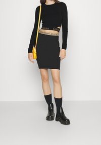 Svart åtsittande långärmad topp med utskärning, tillsammans med en svart knälång kjol med en midjeband från Calvin Klein. Gul axelväska och svarta stövlar.