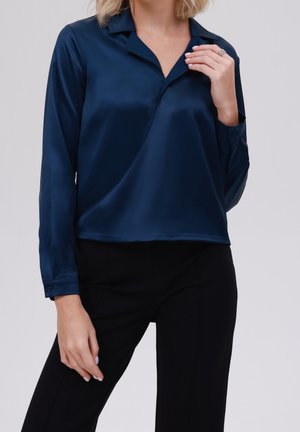 Femme portant un chemisier en satin bleu marine à manches longues avec un col, associé à un pantalon noir, main près du col montrant une bague.