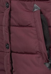 Tejido de chaqueta acolchada burdeos, diseño acolchado con una textura suave, que cuenta con un bolsillo con cremallera y un acento negro cerca de la costura.