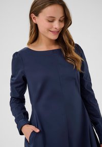 Tamsiai mėlynas suknelė su ilgomis rankovėmis, apvaliu iškirpimu ir klostuota sijonu. Yra šoninės kišenės ir surišimo detalės rankovių kraštuose.