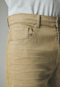 Beige denimbukser med lige snit, der har en frontlomme, bæltestropper og synlige syningsdetaljer. Blød tekstur, slim fit.