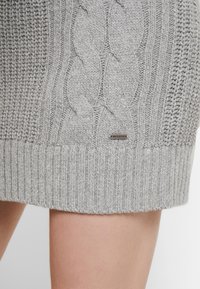 Lätt grå kabelstickad tröja med ribbad fåll och mjuk textur. Logotavla fäst nära fållen ger en subtil accent.