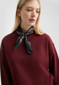 Felpa bordeaux con finitura testurizzata, caratterizzata da una sciarpa nera a fantasia annodata al collo, che aggiunge contrasto e interesse visivo.
