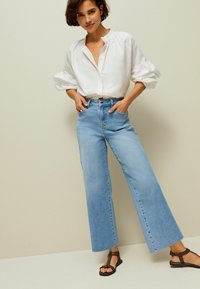 Camisa de algodón blanca con mangas abullonadas, combinada con jeans de cintura alta de pierna ancha en un tono azul claro y sandalias marrones, sobre un fondo neutro.