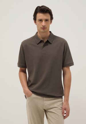 Jonge man met donker haar, gekleed in een bruin poloshirt met korte mouwen en beige broek, staand met één hand in de zak tegen een effen achtergrond.