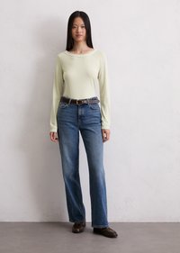Lys grønn langermet topp med bred halsåpning, kombinert med blå rette jeans og brun belte. Modellen har på seg brune sko.