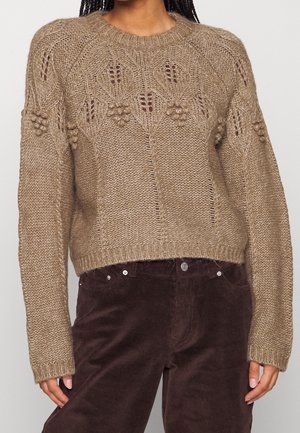 Person trägt einen beigen, gestrickten Pullover mit Blatt- und Bommelmustern, kombiniert mit dunkelbraunen Cordhosen, steht vor einem einfarbigen Hintergrund.