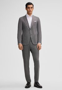 Completo grigio su misura con un tessuto strutturato. Giacca monopetto, rever a punta, camicia bianca sotto e scarpe nere.