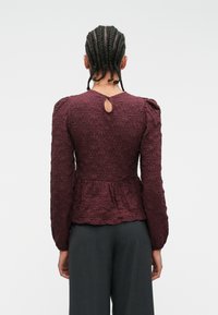 Blusa bordeaux strutturata con maniche lunghe, spalle a palloncino e dettaglio a chiave sul retro, abbinata a pantaloni grigi a vita alta.