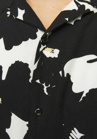 Sort og hvid kortærmet skjorte med et markant blomstermønster, spids krave og en enkelt sort knaplukning ved halsen.