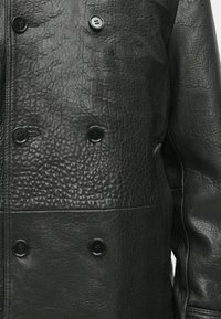 Zwarte leren jas met een gestructureerde afwerking, voorzien van een dubbele rij knopen en vier ronde zwarte knopen.