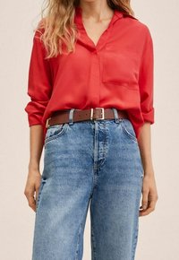 Donna che indossa una blusa rossa a maniche lunghe inserita in jeans blu a vita alta, con una cintura in pelle marrone e fibbia in metallo.