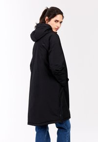 Sort lang kåbe med hætte, to sidelommer, vandafvisende stof og en lige snitdesign. Har subtil branding på skulderen.