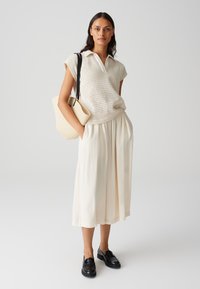 Polo en tricot beige à manches courtes, associé à une jupe ample crème. Accentué par un sac texturé beige et des mocassins noirs.
