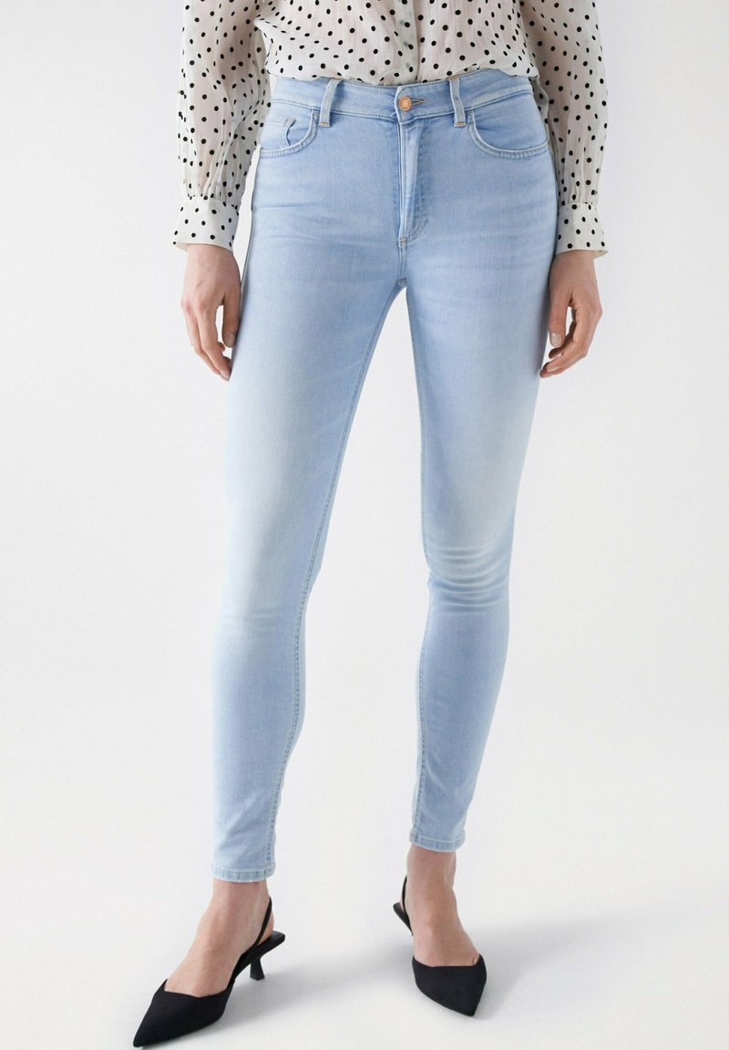 Jeans ajustados de color azul claro en tejido de mezclilla, con una cintura de altura media, frente plano y bolsillos traseros. Combinados con tacones negros de punta afilada.