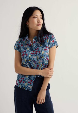 Frau mit langen schwarzen Haaren, die ein blaues kurzärmeliges Blumenshirt und eine dunkle Hose trägt, schaut seitlich und hat die Arme sanft verschränkt.