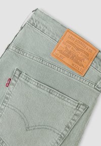 Πίσω τσέπη ανοιχτοπράσινου τζιν Levi's με κόκκινη ετικέτα Levi's και δερμάτινο ταμπελάκι σε ταμπά χρώμα που δείχνει το μέγεθος W32 L32 και τον κωδικό Lot 502.