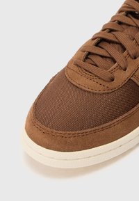 Bruine sneaker van textuur canvas en suède, met een ronde neus en een crèmekleurige rubbersole. Vetersluiting zichtbaar bovenop.