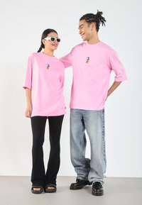 Rosa oversized t-skjorter med trykk av en tegneseriefigur, kombinert med svarte vide bukser og klumpete svarte sko, skaper et avslappet utseende.