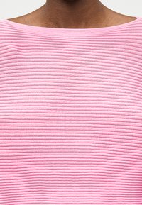Personne à la peau foncée portant un haut en tricot côtelé rose clair avec un décolleté large et arrondi, montré du cou à la partie supérieure de la poitrine.
