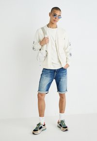 Veste à zip couleur crème clair avec manches imprimées, t-shirt blanc, short en denim et baskets multicolores. Les accessoires comprennent un petit sac et des lunettes de soleil.