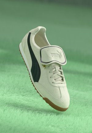 White suede athletic shoe with dark green side stripe, gold logo, lace-up front, and velcro strap. Brown rubber sole.