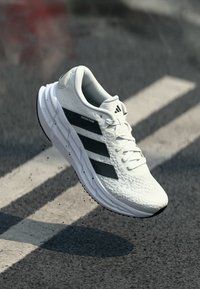 Witte sportieve schoen met een bovenwerk van mesh, zwarte strepen en een gestructureerde outsole. Lichtgewicht ontwerp, met een vetersluiting en een gevoerde kraag.