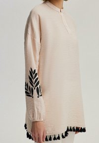 Tunic rosa chiaro con colletto alla cinese, maniche lunghe con ricamo di foglie nero e nappine nere lungo l'orlo. Tessuto testurizzato.