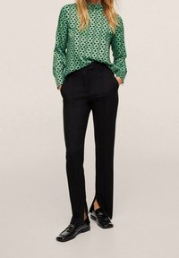 Grüne gemusterte Bluse mit langen Ärmeln, kombiniert mit schwarzen, taillierten Hosen mit Schlitzen an den Knöcheln und schwarzen Loafers.