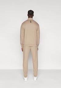 Bēša sporta tērpa komplekts ar rāvējslēdzēju aizdares jaku ar augstu apkakli un smalku detaļu aizmugurē, kopā ar līdzīgi nokarenām biksēm un baltām kurpēm.