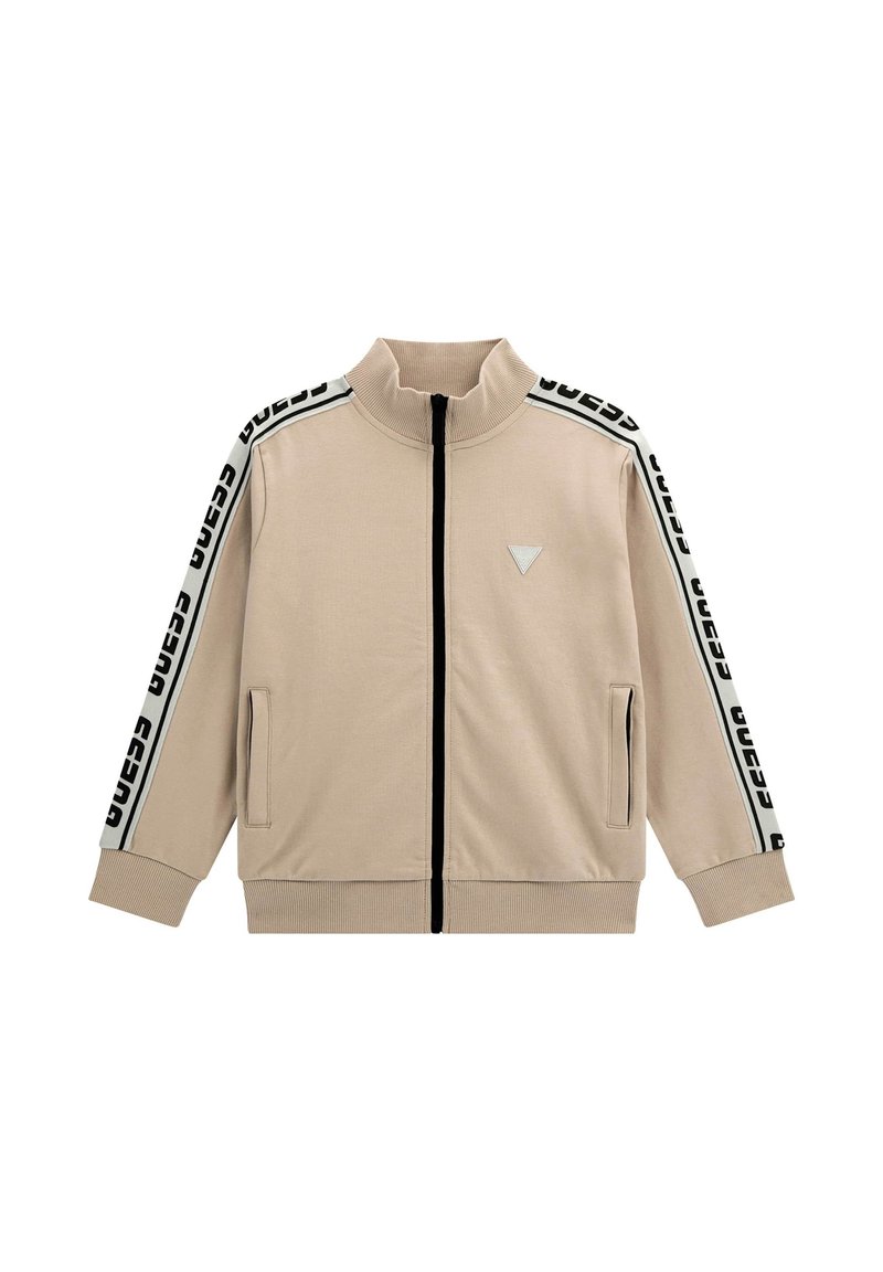 Veste beige zippée avec col haut, fermeture éclair noire, poches latérales et rayures noires contrastantes avec des lettres blanches le long des manches.