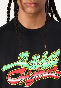 Black t-shirt featuring bold, multicolored "Adidas Originals" graphic in green, red, and yellow. Short sleeves and a round neckline.