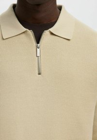 Beżowy sweter na drutach z kołnierzem na zamek i teksturowanym splotem. Posiada subtelne prążkowane detale oraz metalowy zamek.