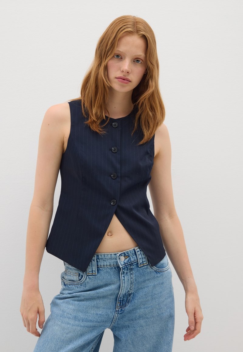 Gilet sans manches navy à rayures verticales, avec cinq grands boutons noirs et un ourlet en pointe, associé à un jean bleu clair.