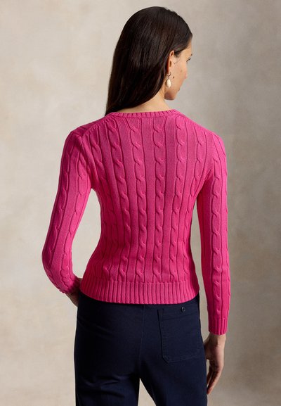 Vrouw met lang donker haar, gekleed in een felroze kabelgebreide trui en donkere broek, met haar rug naar de camera tegen een neutrale achtergrond.