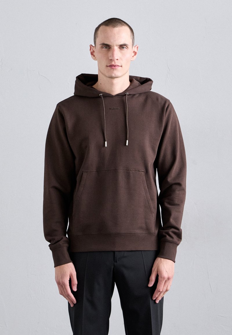 Felpa con cappuccio marrone, dotata di tasca anteriore, stringhe regolabili e polsini a costine; indossata con pantaloni neri su uno sfondo neutro.