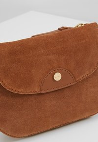 Bruine suède pouch met een afgeronde klep, voorzien van een goudkleurige druksluiting en subtiele stikseldetails langs de randen.