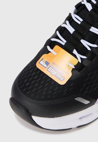 Close-up van een zwarte Skechers instapschoen met witte elastische veters en een oranje label dat het handsfree instapontwerp aangeeft.