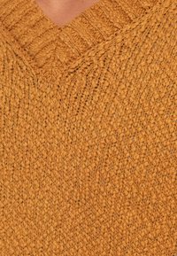 Κοντινή λήψη υφασμένου μουσταρδί κίτρινου πουλόβερ με V λαιμόκοψη, που δείχνει περίπλοκους καναλιούς και σχέδια ραβδώσεων.