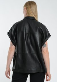 Sort læder oversized skjorte med korte ærmer, der har en glat overflade og minimal syning synlig på ryggen.