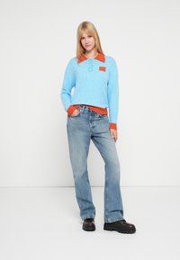 Hellblauer Pullover mit orangefarbenem Kragen und Taschen-Detail, kombiniert mit ausgestellten blauen Jeans und klobigen schwarzen Plateauschuhen.