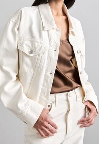 Bílá džínová bunda s volnějším střihem, vybavená dvěma předními kapsami, stříbrnými knoflíky a strukturovaným límcem. Nasažena přes hnědé tričko a bílé kalhoty.