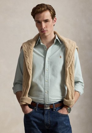 Ung mand med lyseblå skjorte med knapper, beige kabelstrikket sweater lagt over skuldrene, blå jeans, brun bælte og ur på håndleddet, stående med hænderne i lommerne.