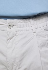 Pantaloni in cotone grigio chiaro con vita elasticizzata, davanti piatto e chiusura con bottone. Sopra i pantaloni è sovrapposta una maglia azzurro pallido.