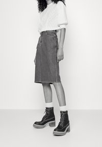 Mujer vistiendo un suéter blanco texturizado, una falda de mezclilla hasta la rodilla, calcetines blancos y botas de plataforma negras con cordones y detalles de cadenas.