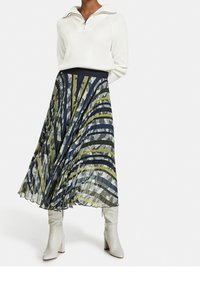 Vihreän, sinisen ja harmaan monivärisellä raidallisella kuviolla oleva laskostettu midi-hame, yhdistettynä valkoiseen vetoketjulliseen neulepaitaan ja valkoisiin korollisiin nilkkureihin.