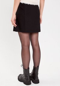 Frau, die schwarze Knöchelstiefel, durchsichtige schwarze Strumpfhosen und einen schwarzen Minirock trägt und vor einem weißen Hintergrund steht.