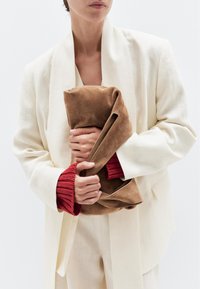 Blazer di lino beige con ampi rever, polsini a maglia rossi e una borsa in suede marrone tenuta strettamente. Texture liscia e design semplice ed elegante.